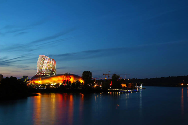 The wine decanter shaped La Cité du Vin lit up at dusk with a light show in orange and reds in Bordeaux, France