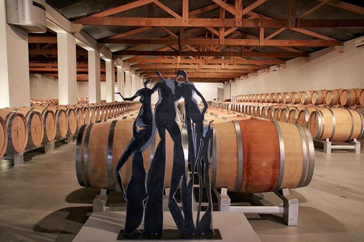 A metal sculpture of three women stands in front of the rows of barrels at Château Marquis de Terme