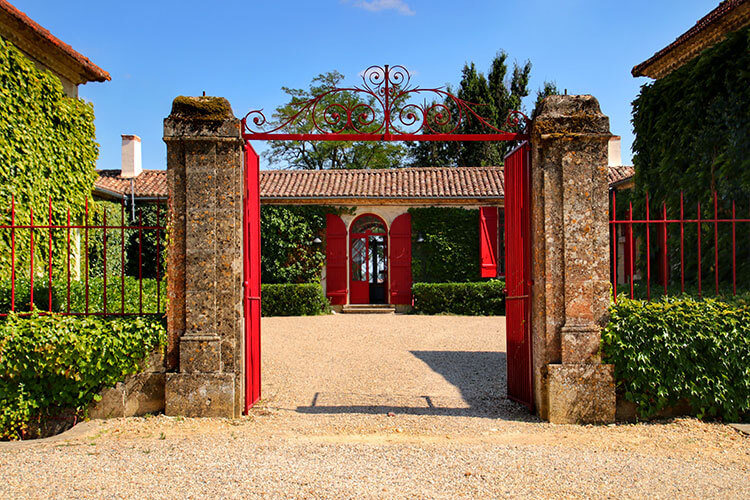 A red gate frames the single level former stone farmhouse with red doors and shutters at Chateau Sigalas Rabaud