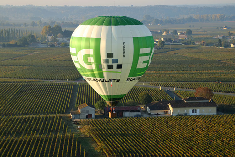 The green and white Girone hot air balloon flying low over a château and vineyards in Saint-Émilion