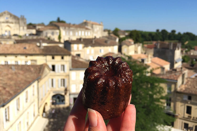 Jennifer holds up a canelé in Saint-Émilion with the village blurred behind it