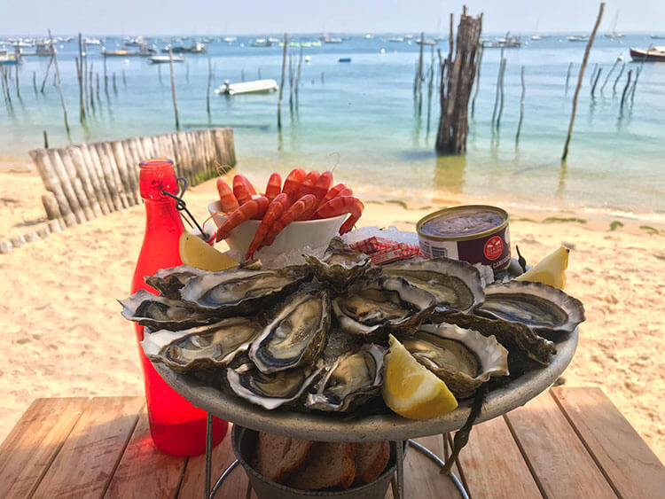 A plate of a dozen oysters on the table with the beach and Bassin of Arcachon behind at Emile et un Huîte in Village l'Herbe on Cap Ferret