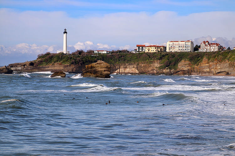 More than a dozen surfers paddle out in the waves at Grande Plage in Biarritz with the lighthouse looming in the distance