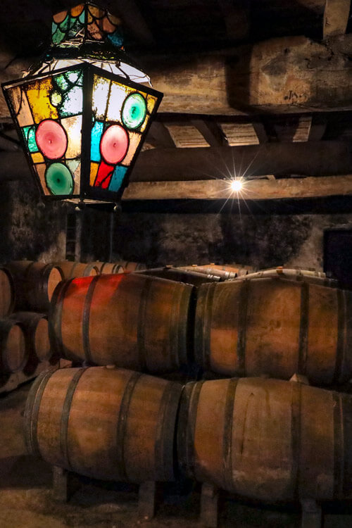 A glass bottle lamp from South America hangs in the historic barrel room casting multicolored light in shades od pink, green and yellow on the barrels of aging Cérons wine at Château de Cérons