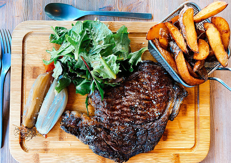 A cut of entrecôte beef served on a cutting board with fries in a little basket at Atelier de Candale