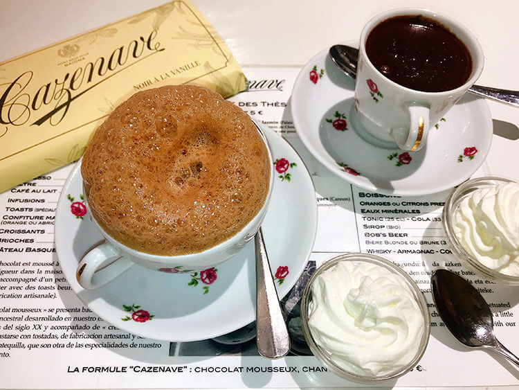 A bar of chocolate, a cup of hot chocolate with a foamy top and a cup of thick pudding-like hot chocolate served with whipped cream