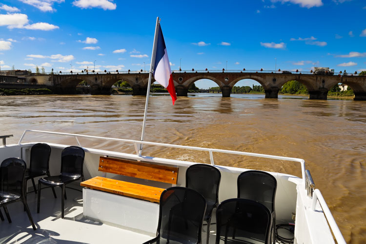 A French flag flutters from the back of the boat with the stone bridge of Libourne behind