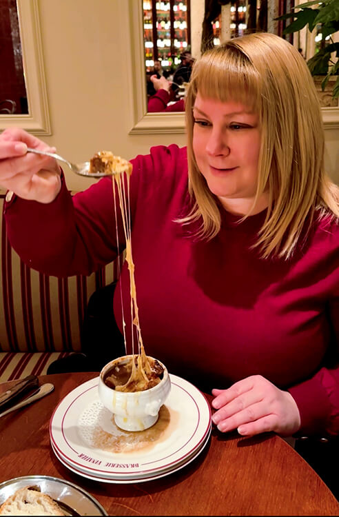 Jennifer lifts a spoon high up with the melted cheese stretching from the cup of onion soup at Brasserie Vinatier